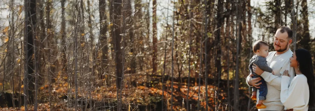 Proud mother and father both wearing white sweaters holding their baby who is wearing a grey sweater in a forest setting