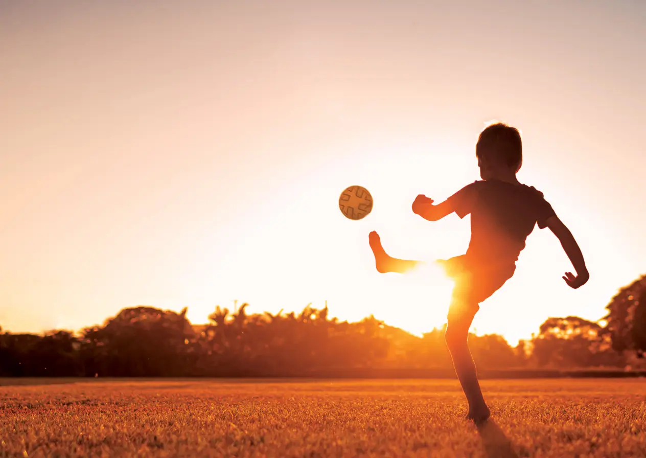 child kicking a soccer ball in a field with the sun setting over the trees
