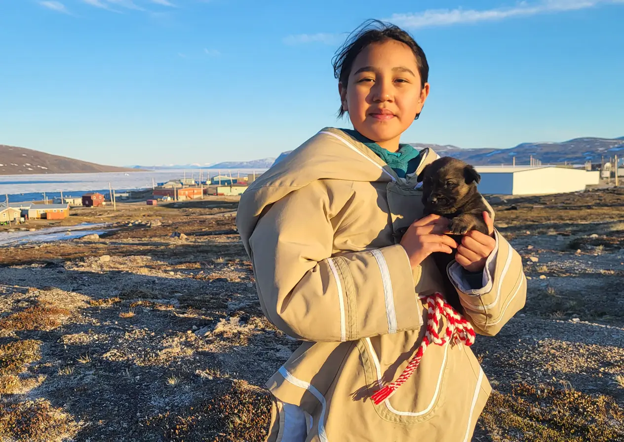 A teenage girl wearing a traditional amauti holding a puppy. She is standing on a tundra field with houses behind.