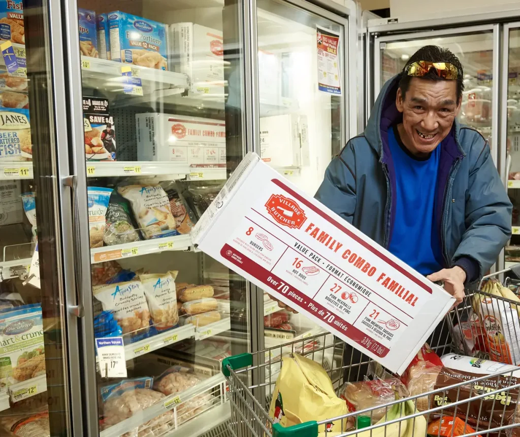 indigenous older man standing in front of a store freezer holding a Family Combo box of meat.