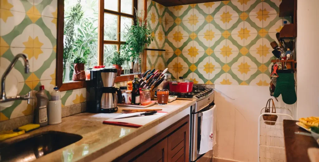 A homey kitchen counter with a sink and a coffee machine and a dishwasher, a nice pattern on the wall with a window over the counter