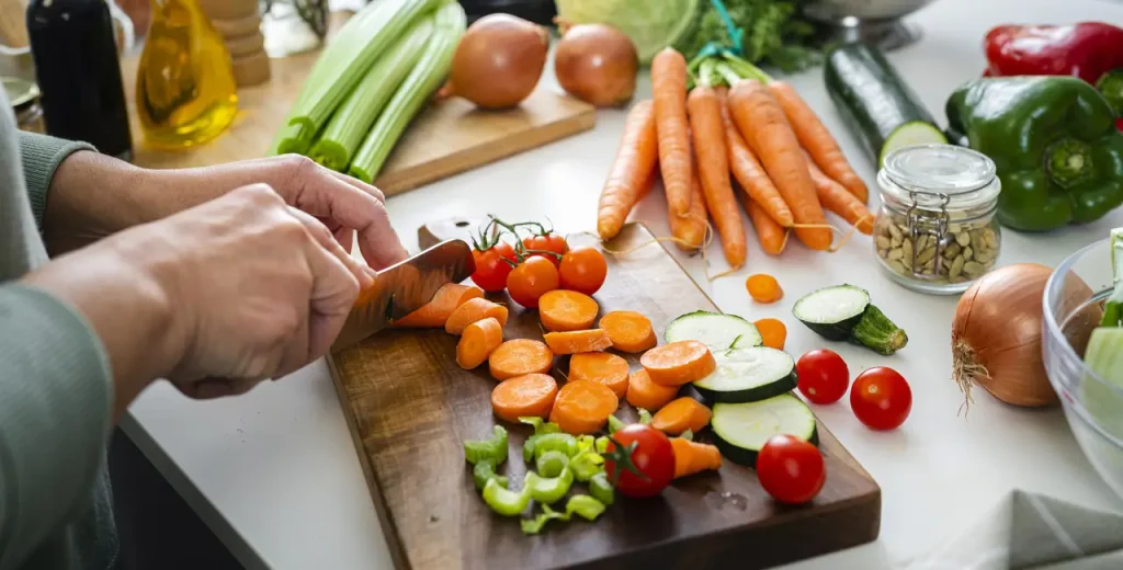 Female hands cutting up carrots, celery, cucumber on a cutting board with other veggies nearby