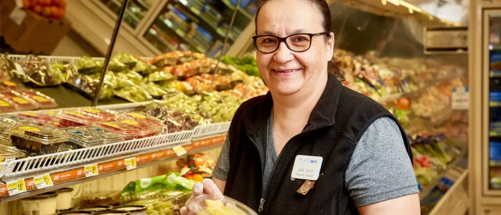 Older employee wearing plastic gloves standing in front of the produce container holding a package of fruit