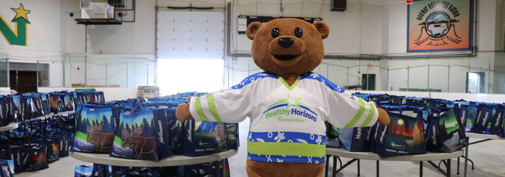 HHF mascot Northy who is a brown bear wearing a hockey jersy with the HHF logo on it is standing if front of hamper bags for the community