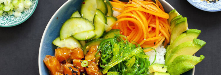 a bowl full of carrots, avocados, cucumbers in a blue bowl sitting on a dark table shot from above