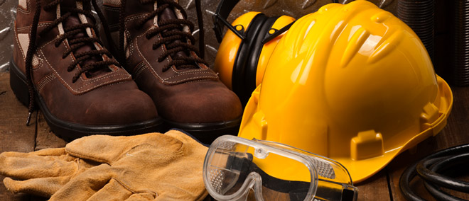 Yellow hard-hat with safety goggles. Work gloves laying on the ground with work boots in the back along with ear protection